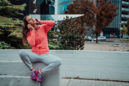 Téléchargez les photos : Une femme en tenue de sport se repose dans un environnement urbain après une séance d'entraînement matinale difficile tout en utilisant des écouteurs silencieux. - en image libre de droit