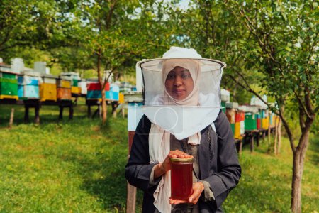 Foto de Portrait of a Muslim African American woman in the beekeeping department of a honey farm holding a jar of honey in her hand. - Imagen libre de derechos