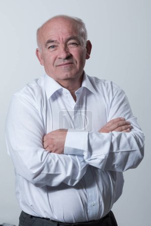 Téléchargez les photos : Confident senior man in white shirt crossing hands on chest and looking at camera while standing against gray background. Self confident senior isolated white studio shoot. High quality photo - en image libre de droit