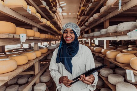 Téléchargez les photos : Femme d'affaires musulmane afro-américaine vérifiant la qualité des produits et entrant des données dans un ordinateur portable dans une entreprise locale de fabrication de fromage. - en image libre de droit