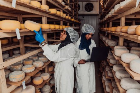 Téléchargez les photos : Entreprise d'un partenaire musulman dans un entrepôt de fromage, contrôle de la qualité du fromage et saisie de données dans un ordinateur portable. - en image libre de droit