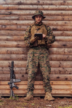 Foto de Soldado usando tableta contra vieja pared de madera en el campamento militar para mantenerse en contacto con amigos y familiares. - Imagen libre de derechos