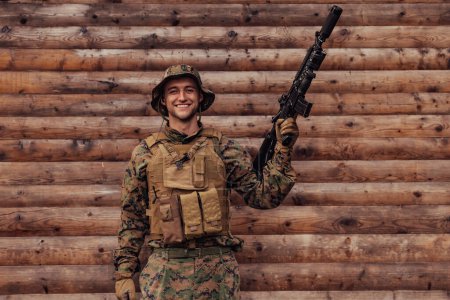Foto de Un soldado de uniforme con un rifle en la mano está parado frente a una pared de madera. Un soldado protege la base forestal del enemigo. - Imagen libre de derechos