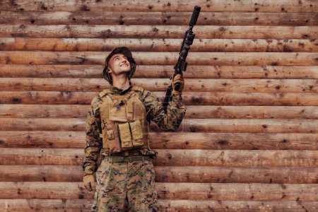 Photo for A soldier in uniform with a rifle in his hand is standing in front of a wooden wall. A soldier guards the forest base from the enemy. - Royalty Free Image