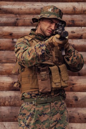 Téléchargez les photos : Un soldat en uniforme avec un fusil à la main se tient devant un mur de bois. Un soldat protège la base forestière de l'ennemi. - en image libre de droit
