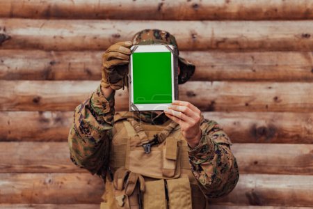 Téléchargez les photos : Soldat utilisant un ordinateur tablette contre un vieux mur en bois dans un camp militaire pour rester en contact avec ses amis et sa famille. - en image libre de droit