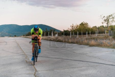 Photo for Triathlete riding his bicycle during sunset, preparing for a marathon. The warm colors of the sky provide a beautiful backdrop for his determined and focused effort - Royalty Free Image