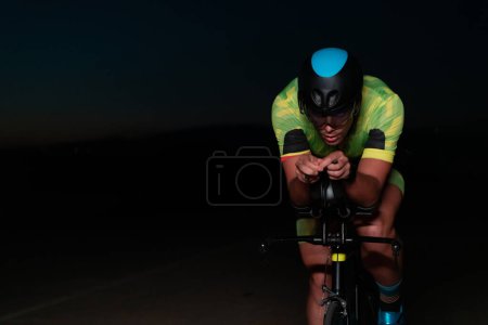 Téléchargez les photos : Un triathlète fait du vélo dans l'obscurité de la nuit, se poussant à se préparer pour un marathon. Le contraste entre l'obscurité et la lumière de son vélo crée un sentiment de drame et met en évidence le - en image libre de droit