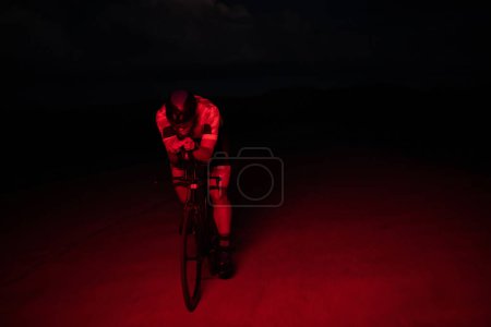 Foto de Un triatleta monta su bicicleta en la oscuridad de la noche, empujándose para prepararse para un maratón. El contraste entre la oscuridad y la luz de su moto crea una sensación de drama y destaca la - Imagen libre de derechos