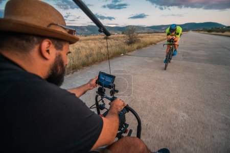 Photo for A videographer recording a triathlete riding his bike preparing for an upcoming marathon.Athletes physical endurance and the dedication required to succeed in the sport - Royalty Free Image