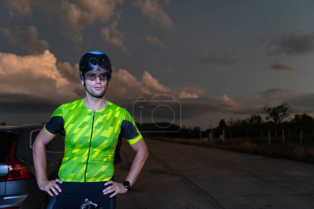 Foto de Un triatleta descansando en la carretera después de un duro paseo en bicicleta en la noche oscura, apoyado en su bicicleta en completo agotamiento. - Imagen libre de derechos