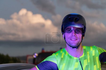 Foto de Un triatleta descansando en la carretera después de un duro paseo en bicicleta en la noche oscura, apoyado en su bicicleta en completo agotamiento. - Imagen libre de derechos