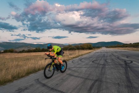 Photo for Triathlete riding his bicycle during sunset, preparing for a marathon. The warm colors of the sky provide a beautiful backdrop for his determined and focused effort - Royalty Free Image