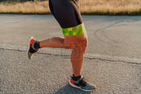 Photo for Closeup of muscular legs of a triathlete in professional equipment running early in the morning, preparing for a marathon, dedication to sports and readiness for marathon challenges. - Royalty Free Image