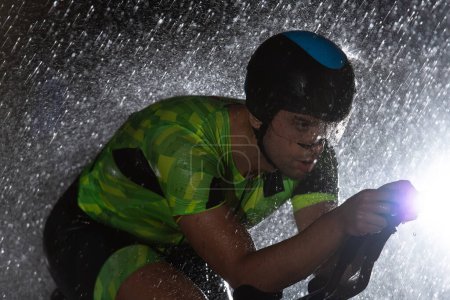 Téléchargez les photos : Un triathlète bravant la pluie alors qu'il roule toute la nuit, se préparant pour le prochain marathon. Les gouttes de pluie floues au premier plan et l'atmosphère sombre et humide en arrière-plan ajoutent - en image libre de droit