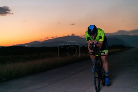 Téléchargez les photos : Un triathlète fait du vélo dans l'obscurité de la nuit, se poussant à se préparer pour un marathon. Le contraste entre l'obscurité et la lumière de son vélo crée un sentiment de drame et met en évidence le - en image libre de droit