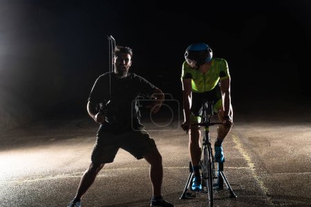 Foto de Entre bastidores. Videógrafo profesional en una sesión nocturna con un triatleta montando una bicicleta para grabar videos. - Imagen libre de derechos