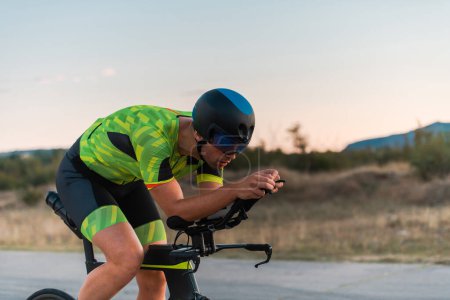 Téléchargez les photos : Triathlète en vélo pendant le coucher du soleil, se préparant pour un marathon. Les couleurs chaudes du ciel fournissent une belle toile de fond pour son effort déterminé et concentré - en image libre de droit