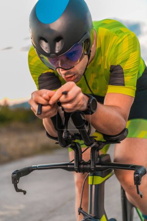 Foto de Foto de cerca del triatleta montando su bicicleta durante el atardecer, preparándose para un maratón. Los colores cálidos del cielo proporcionan un hermoso telón de fondo para su esfuerzo decidido y centrado - Imagen libre de derechos