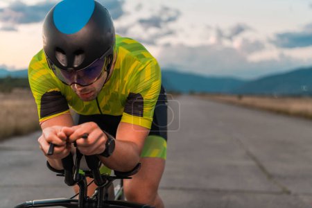 Foto de Foto de cerca del triatleta montando su bicicleta durante el atardecer, preparándose para un maratón. Los colores cálidos del cielo proporcionan un hermoso telón de fondo para su esfuerzo decidido y centrado - Imagen libre de derechos