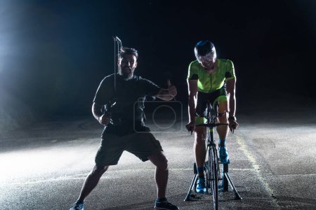 Foto de Entre bastidores. Videógrafo profesional en una sesión nocturna con un triatleta montando una bicicleta para grabar videos. - Imagen libre de derechos