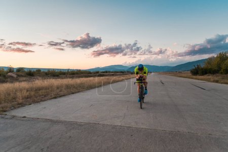 Téléchargez les photos : Triathlète en vélo pendant le coucher du soleil, se préparant pour un marathon. Les couleurs chaudes du ciel fournissent une belle toile de fond pour son effort déterminé et concentré - en image libre de droit
