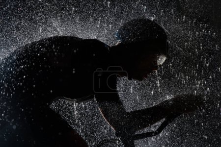 Photo for A triathlete braving the rain as he cycles through the night, preparing himself for the upcoming marathon. The blurred raindrops in the foreground and the dark, moody atmosphere in the background add - Royalty Free Image