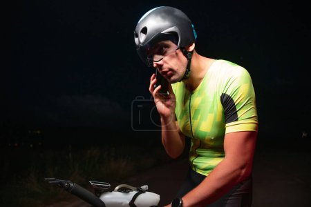 Téléchargez les photos : Un triathlète utilisant un smartphone tout en prenant une pause d'un entraînement de cyclisme de nuits difficiles. Photo de haute qualité - en image libre de droit
