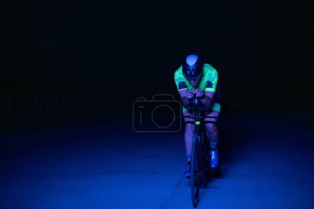 Foto de Un triatleta monta su bicicleta en la oscuridad de la noche, empujándose para prepararse para un maratón. El contraste entre la oscuridad y la luz de su moto crea una sensación de drama y destaca la - Imagen libre de derechos