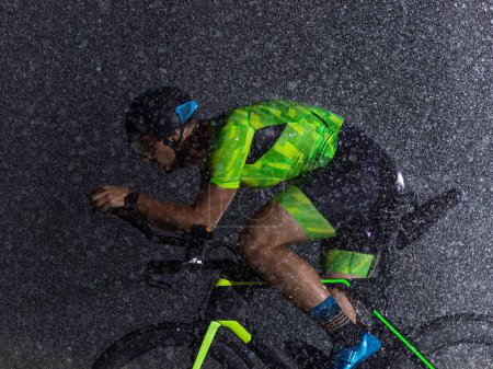 Téléchargez les photos : Un triathlète bravant la pluie alors qu'il roule toute la nuit, se préparant pour le prochain marathon. Les gouttes de pluie floues au premier plan et l'atmosphère sombre et humide en arrière-plan ajoutent - en image libre de droit