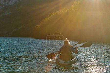 Photo for A young couple enjoying an idyllic kayak ride in the middle of a beautiful river surrounded by forest greenery in sunset time. - Royalty Free Image
