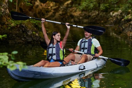 Photo for A young couple enjoying an idyllic kayak ride in the middle of a beautiful river surrounded by forest greenery. - Royalty Free Image