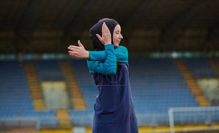 Téléchargez les photos : Une femme musulmane dans une burqa, une tenue de sport islamique, fait des exercices corporels, étirant son cou, ses jambes et son dos après une séance d'entraînement difficile sur le parcours du marathon - en image libre de droit