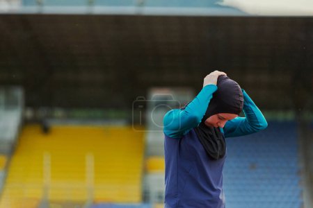 Photo for A Muslim woman in a burqa, an Islamic sports outfit, is doing body exercises, stretching her neck, legs and back after a hard training session on the marathon course - Royalty Free Image