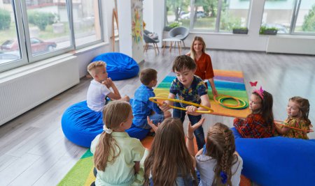 Téléchargez les photos : Une enseignante heureuse assise et jouant à des jeux de mains avec un groupe de petits écoliers. - en image libre de droit