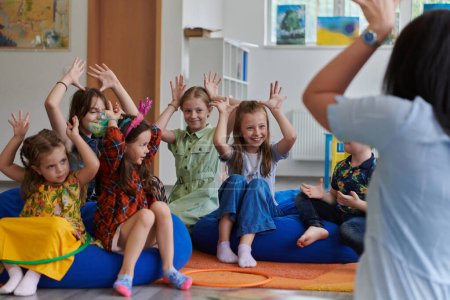 Téléchargez les photos : Une enseignante heureuse assise et jouant à des jeux de mains avec un groupe de petits écoliers. - en image libre de droit