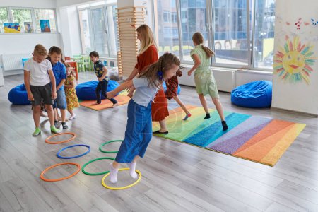 Foto de Pequeños niños de guardería con maestra en el suelo en el aula, haciendo ejercicio. Saltar sobre la pista círculos hula hoop en el suelo - Imagen libre de derechos