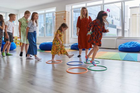 Photo for Small nursery school children with female teacher on floor indoors in classroom, doing exercise. Jumping over hula hoop circles track on the floor - Royalty Free Image
