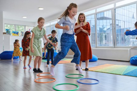 Foto de Pequeños niños de guardería con maestra en el suelo en el aula, haciendo ejercicio. Saltar sobre la pista círculos hula hoop en el suelo - Imagen libre de derechos