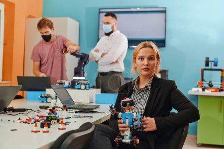 Foto de Una mujer sentada en un laboratorio y resolviendo problemas y analizando la verificación de robots. En segundo plano, los colegas están hablando en una reunión en línea. - Imagen libre de derechos