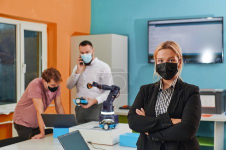 Foto de Una mujer con una máscara protectora de pie en un laboratorio mientras sus colegas prueban un nuevo invento robótico en el fondo - Imagen libre de derechos