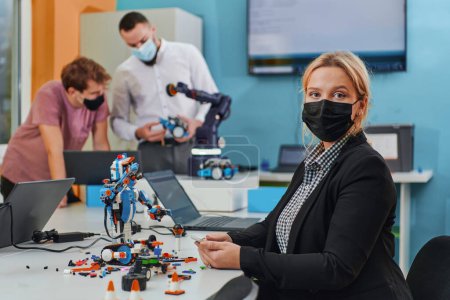 Foto de Una mujer con una máscara protectora de pie en un laboratorio mientras sus colegas prueban un nuevo invento robótico en el fondo - Imagen libre de derechos
