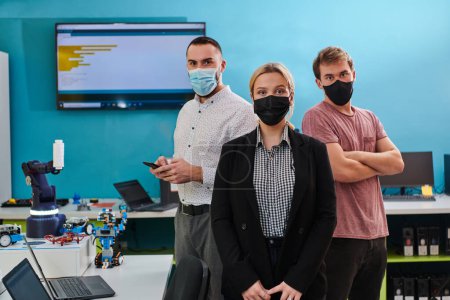 Téléchargez les photos : Un groupe de collègues se tient debout dans un laboratoire de robotique, les bras croisés, portant des masques de protection, symbolisant leur travail d "équipe et leur engagement envers l'innovation technologique et la recherche scientifique - en image libre de droit
