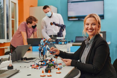 Foto de Una mujer sentada en un laboratorio y resolviendo problemas y analizando la verificación de robots. En segundo plano, los colegas están hablando en una reunión en línea. - Imagen libre de derechos
