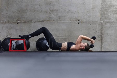 Photo for A fit woman is lying on the gym floor, performing arm exercises with dumbbells and showcasing her dedication and strength - Royalty Free Image