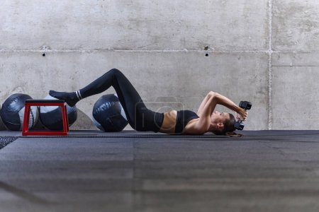 Téléchargez les photos : Une femme en forme est allongée sur le sol de la salle de gym, effectuant des exercices de bras avec des haltères et montrant son dévouement et sa force - en image libre de droit
