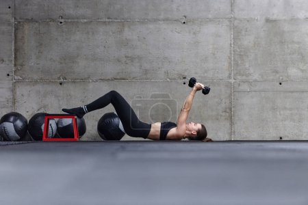Foto de Una mujer en forma está tumbada en el suelo del gimnasio, realizando ejercicios de brazo con pesas y mostrando su dedicación y fuerza - Imagen libre de derechos