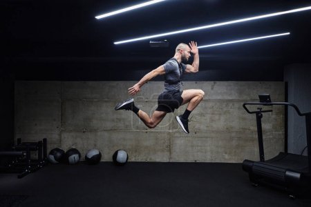 Foto de Un hombre musculoso capturado en el aire mientras salta en un gimnasio moderno, mostrando su atletismo, poder y determinación a través de una rutina de fitness de alta intensidad. - Imagen libre de derechos