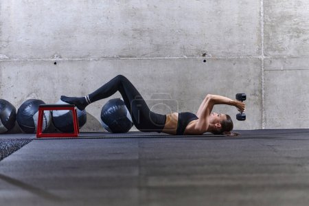Foto de Una mujer en forma está tumbada en el suelo del gimnasio, realizando ejercicios de brazo con pesas y mostrando su dedicación y fuerza - Imagen libre de derechos