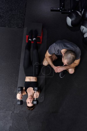Téléchargez les photos : Un homme musclé aidant une femme en forme dans une salle de gym moderne alors qu'ils s'engagent dans divers exercices du corps et des étirements musculaires, montrant leur dévouement à la forme physique et bénéficiant du travail d'équipe et de soutien. - en image libre de droit